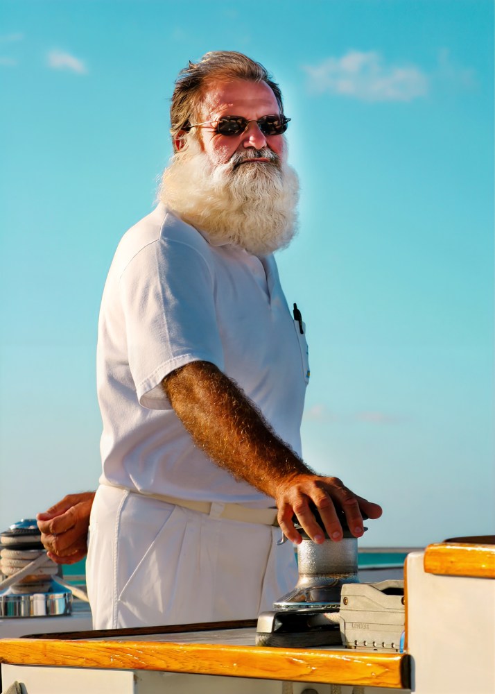 Man with white beard and sunglasses steering a boat under a clear blue sky.