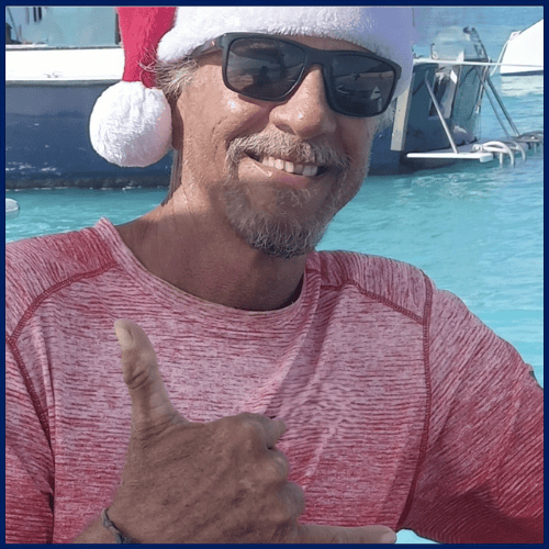 Man in Santa hat and sunglasses smiles, giving a shaka sign by the water.