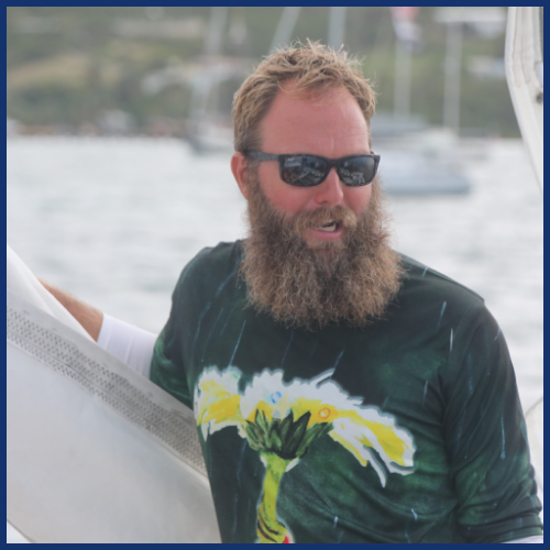 Bearded man in sunglasses on a sailboat, wearing a tropical-themed shirt.