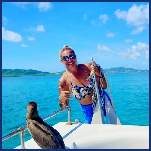 Person in floral top on boat smiles at bird on railing, with blue ocean and islands in background.
