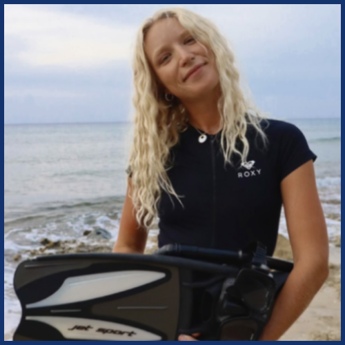 Person with long blonde hair holds snorkeling gear by the beach.