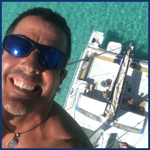 Man wearing sunglasses smiles on a sailing boat with turquoise water below.