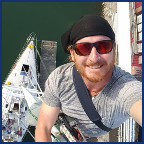 Man with red beard taking selfie on a sailboat mast, wearing sunglasses and a black headscarf.