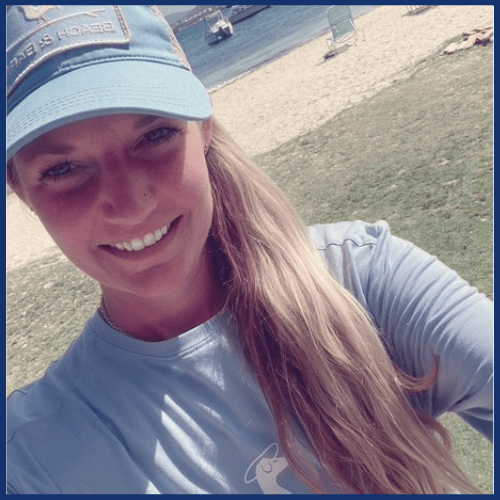 Person in a blue cap and shirt smiling on a beach with water in the background.
