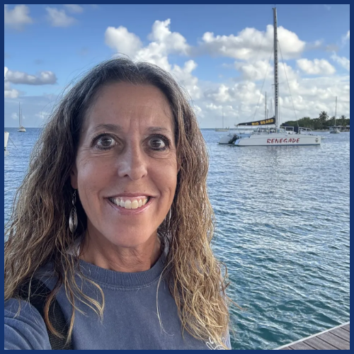 Woman smiling by the sea with sailboats in the background on a sunny day.